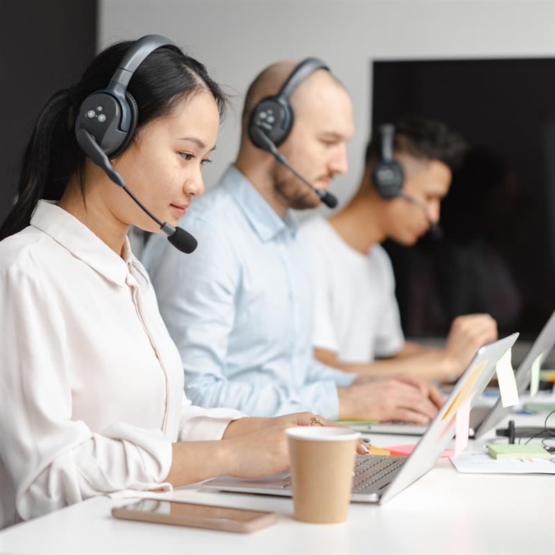 Three people sitting at a desk with headsets on and working on their laptops.